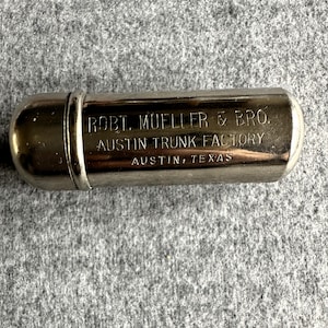 Puede incluir: Un tubo de metal plateado con el texto "ROBT. MUELLER & BRO. AUSTIN TRUNK FACTORY AUSTIN, TEXAS" grabado en el lateral.