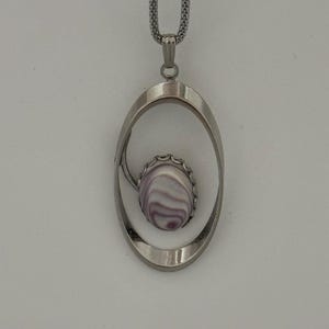 May include: A silver-toned pendant necklace featuring an oval frame with a polished finish. Centered within the frame is a small, oval-shaped stone with concentric purple and white swirling patterns, suspended from a matching silver-toned mesh chain.