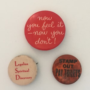 Puede incluir: Tres botones redondos con diferentes mensajes. El botón superior es rojo con texto blanco que dice "now you feel it - now you don't!" El botón izquierdo es blanco con texto rojo que dice "Legalize Spiritual Discovery". El botón derecho es naranja con texto negro que dice "STAMP OUT PAY TOILETS".