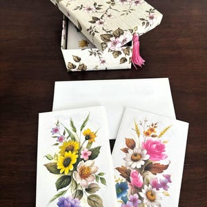 May include: A floral stationery set featuring a cream-colored box with a pink tassel and a matching floral design. The set includes white cards adorned with colorful flower bouquets, including sunflowers, roses, and daisies.