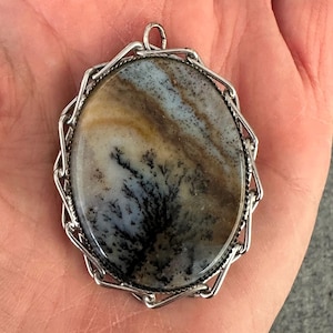 May include: An oval-shaped pendant featuring a polished dendritic agate stone with brown, tan, and white bands and black fern-like inclusions. The stone is set in a silver-toned metal frame with a decorative, geometric, chain-link style border.