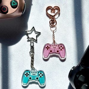May include: Two acrylic keychains shaped like game controllers. The keychain on the left is blue and says "Player One". The keychain on the right is pink and says "Player Two".