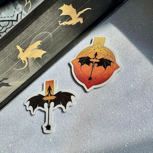 Puede incluir: Dos marcadores con temática de dragones y un libro con ilustraciones de dragones dorados. Los marcadores presentan una silueta de dragón negro sobre un degradado de puesta de sol. El libro tiene diseños de dragones dorados en el lomo.