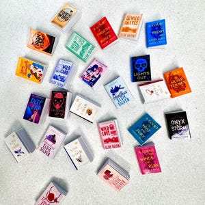 Peut inclure: Une collection de petits livres aux couvertures colorées, avec des titres tels que "Fourth Wing" et "A Court of Thorns and Roses". Les livres sont disposés sur une surface blanche, présentant des romans de fantasy et de romance. On peut voir des livres de Sarah J. Maas.