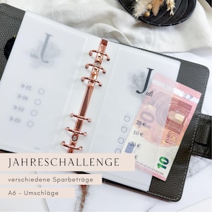 Könnte beinhalten: Ein dunkelgrauer A6-Ordner mit einem roségoldenen Ringmechanismus, der transparente Einsätze enthält. Ein Zehn-Euro-Schein ist sichtbar. Der Text "JAHRESCHALLENGE" und "verschiedene Sparbeträge" sind zu sehen.