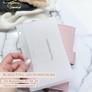 Könnte beinhalten: Ein rosa Planer mit einem transparenten, mattierten Kunststoffeinsatz mit der Aufschrift "LEBENSMITTEL". Der Einsatz wird vor dem Planer gehalten, mit dem Text "BUDGETING GELDMUSCHLAG" und anderem Text unten. Der Planer liegt auf einer Marmoroberfläche.