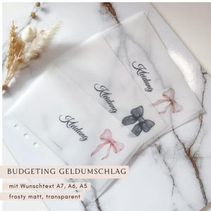 Könnte beinhalten: Transparente, mattierte Budget-Umschläge mit dem Aufdruck "Kleidung". Jeder Umschlag hat eine Aquarellschleife in Rosa oder Grau. Der Text "BUDGETING GELDUMSCHLAG" ist unten aufgedruckt.