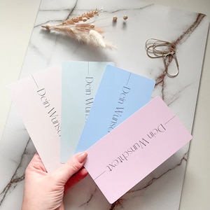 Könnte beinhalten: Vier pastellfarbene Karten, die aufgefächert sind, jede mit dem Text "Dein Wunschtext" in einer minimalistischen Schriftart. Die Karten sind in Rosa-, Blau- und Grüntönen gehalten. Ein Zweig getrockneter Blumen und goldene Büroklammern befinden sich im Hintergrund.