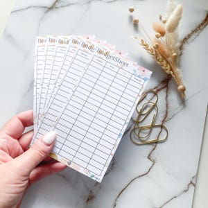 Könnte beinhalten: Ein Stapel Budgetblätter mit floralen Verzierungen und dem Text "Budget Sheet" sowie linierte Abschnitte zur Finanzverfolgung. Goldene Büroklammern und Trockenblumen sind ebenfalls vorhanden.