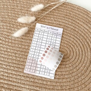 May include: A printable chart for tracking a 50 euro savings goal over 30 days. The chart has columns for day, amount, and points. The chart is printed on white paper and has a pink and grey colour scheme.