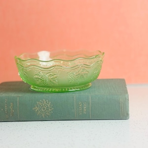 May include: A green glass bowl with a grape design sits atop a vintage green book. The bowl has a scalloped edge and a pedestal base. The book has gold lettering and a sunburst design.