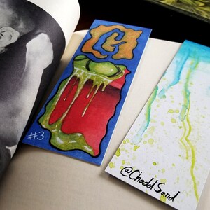 Pu&ograve; includere: Due segnalibri fatti a mano. Un segnalibro presenta un design astratto verde, giallo e rosso con la lettera "G" in arancione. L'altro segnalibro presenta un design acquerello blu e verde con il testo "@Chadd Sand".