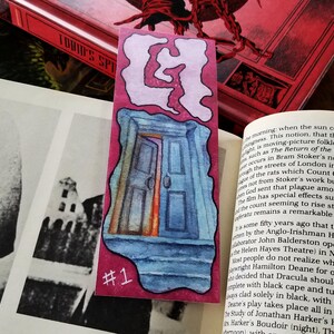 Pu&ograve; includere: Un segnalibro acquerellato rosa e blu con un'illustrazione di una porta e il numero "#1" stampato in basso. Il segnalibro fa parte di un'immagine pi&ugrave; ampia che include un libro e una foto.