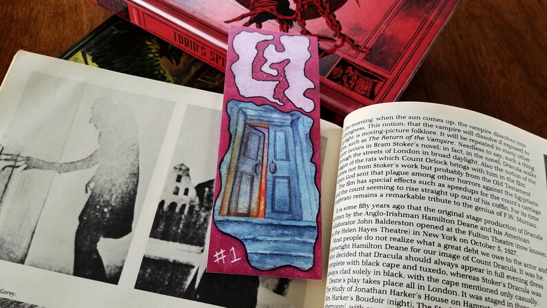 Pu&ograve; includere: Un segnalibro acquerellato rosa e blu con un'illustrazione di una porta e il numero "#1" stampato in basso. Il segnalibro fa parte di un'immagine pi&ugrave; ampia che include un libro e una foto.
