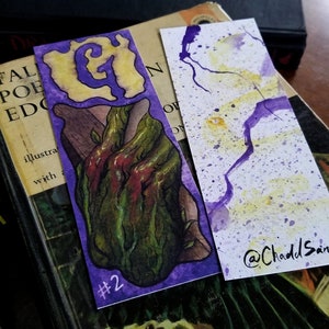 Pu&ograve; includere: Due segnalibri ad acquerello. Il primo segnalibro presenta uno sfondo viola con una lettera G gialla e un'illustrazione verde e rossa di una mano che sporge da una vite. Il secondo segnalibro ha uno sfondo bianco con schizzi di acquerello viola e giallo. Il segnalibro &egrave; firmato "@ChaddSand".