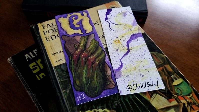 Pu&ograve; includere: Due segnalibri ad acquerello. Il primo segnalibro presenta uno sfondo viola con una lettera G gialla e un'illustrazione verde e rossa di una mano che sporge da una vite. Il secondo segnalibro ha uno sfondo bianco con schizzi di acquerello viola e giallo. Il segnalibro &egrave; firmato "@ChaddSand".
