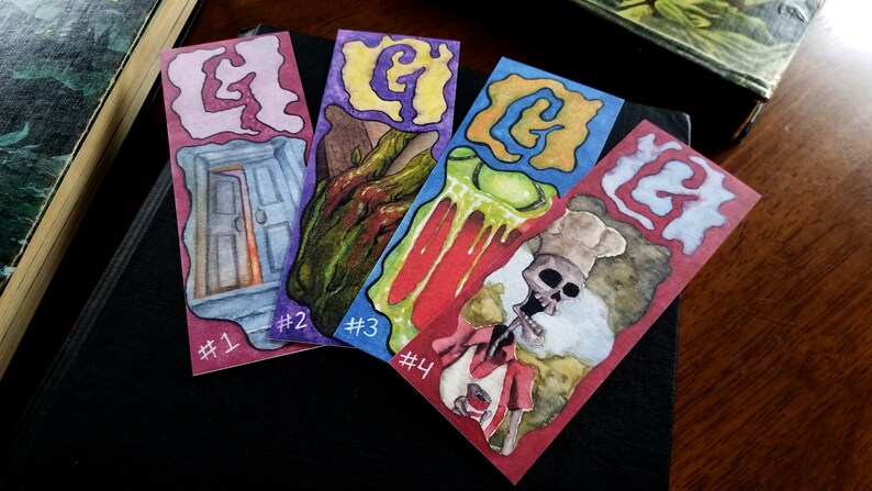 Pu&ograve; includere: Quattro segnalibri ad acquerello con un tema spettrale. Ogni segnalibro presenta un'illustrazione diversa con una lettera "G" al centro. I segnalibri sono numerati da 1 a 4.