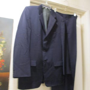 May include: A navy blue suit, including a blazer and trousers, hangs on a hanger. The blazer has a two-button closure and front pockets. The suit is made of a smooth fabric, suitable for formal occasions.