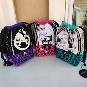 May include: Three black drawstring bags with different colorful animal print fabric bottoms. The bags have different designs on the front, including a skull and crossbones, a cartoon character, and a dog. The bags are all made by the same company, "Evil Twin Designs."