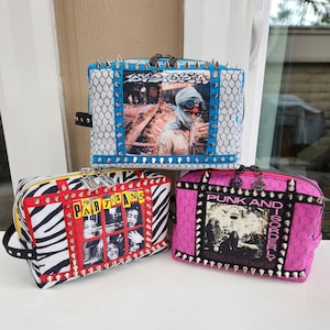 May include: Three zippered cosmetic bags with punk rock band graphics. The bags are black, white, and pink with silver studs. The bags have a zebra print, a white and black honeycomb pattern, and a pink honeycomb pattern. The bags have the band names "The Partisans", "Punk and Disorderly", and "1977".