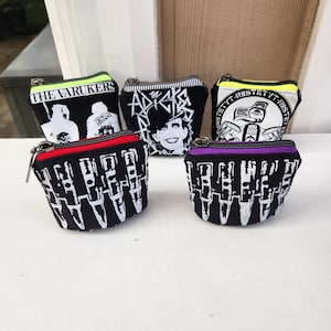 May include: Five small, black zippered pouches with various white graphic designs and text. The pouches have different colored zipper trim: neon green, black, yellow, red, and purple. The text includes "THE VARUKERS" and "THE ADICTS".