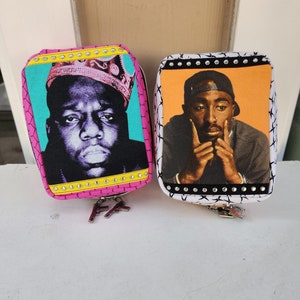 Könnte beinhalten: Zwei kleine Reißverschlusstaschen mit schwarz-weißem Muster. Eine Tasche zeigt ein Porträt von The Notorious B.I.G. mit einer Krone auf dem Kopf. Die andere Tasche zeigt ein Porträt von Tupac Shakur. Beide Taschen haben einen gelben Reißverschluss.