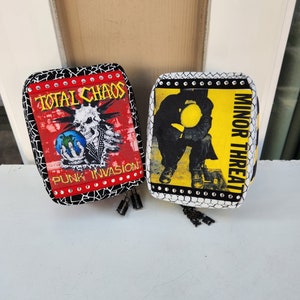 Könnte beinhalten: Zwei kleine, rechteckige Beutel mit schwarz-weiß gemusterten Rändern. Einer hat einen roten Hintergrund mit den Worten "TOTAL CHAOS" und "PUNK INVASION" und einer Totenkopf-Grafik. Der andere hat einen gelben Hintergrund mit den Worten "MINOR THREAT" und einer schwarzen Grafik.