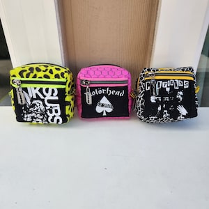 May include: Three small black and white patterned zippered pouches with different designs. The pouch on the left has a yellow and black leopard print background with the text "SUBS" in white. The middle pouch has a pink background with a black honeycomb pattern and the text "Motorhead" in white. The pouch on the right has a black and white graphic print with the text "Citizens" in white.