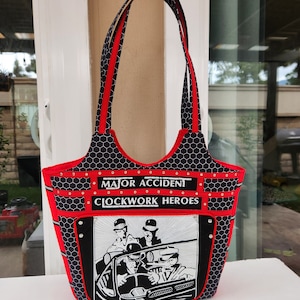Pode incluir: Bolsa tote preta e vermelha. Apresenta um padrão de favo de mel preto, detalhes vermelhos e o texto "MAJOR ACCIDENT CLOCKWORK HEROES". A bolsa tem duas alças vermelhas.