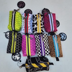 Puede incluir: Nueve pequeñas bolsas con cremallera de diferentes patrones y colores. Las bolsas tienen un logotipo de calavera y huesos cruzados en el tirador de la cremallera. Los patrones incluyen cuadros blanco y negro, estampado de leopardo, estampado de vaca blanco y negro, panal de abeja rosa y negro, panal de abeja amarillo y negro, panal de abeja blanco y negro y un patrón de eslabón de cadena negro y amarillo.
