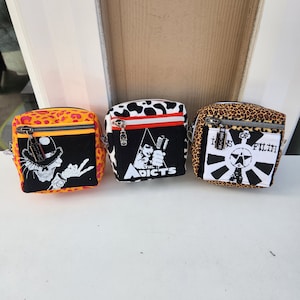 May include: Three small zippered bags with different animal print patterns. The bags have black and white graphic designs with band names and logos. The bags are "The Adicts", "Icons of Filth", and a skull with a top hat.