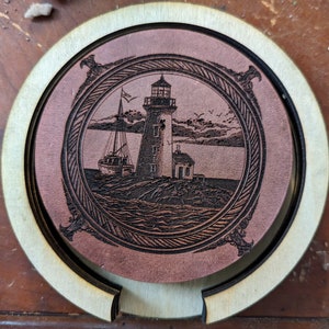 May include: A brown leather coaster with a black and white engraved image of a lighthouse and a boat. The coaster has a wooden outer ring.