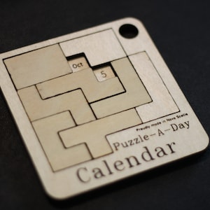 May include: A wooden calendar puzzle with a sliding piece for the date. The puzzle is for October 5th and says "Puzzle-A-Day Calendar" on the bottom.