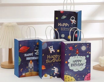 May include: Four blue and white paper gift bags with a space theme. The bags have white handles and feature an astronaut, planets, stars, and rockets. The bags say "Happy Birthday" in gold lettering.
