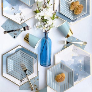 May include: A set of blue and gold paper plates and cups with a watercolour design. The plates are hexagonal and have a white centre with blue and gold stripes. The cups are blue with a gold rim. There are also napkins and forks in the image.