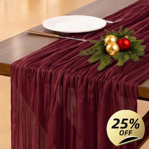 Puede incluir: Camino de mesa burdeos con textura plisada, colocado sobre una mesa de madera. Un plato blanco, un cuchillo y un arreglo festivo de adornos dorados y rojos con vegetación se colocan sobre el camino de mesa. Una pegatina dorada con "25% OFF" está en la esquina inferior derecha.