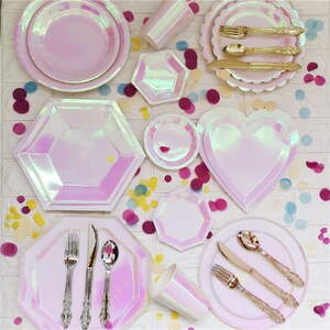 Può includere: Un set di piatti e bicchieri di carta rosa iridescente con bordo smerlato. I piatti sono di varie forme, tra cui rotonda, a forma di cuore e ottagonale. Ci sono anche forchette, coltelli e cucchiai dorati.