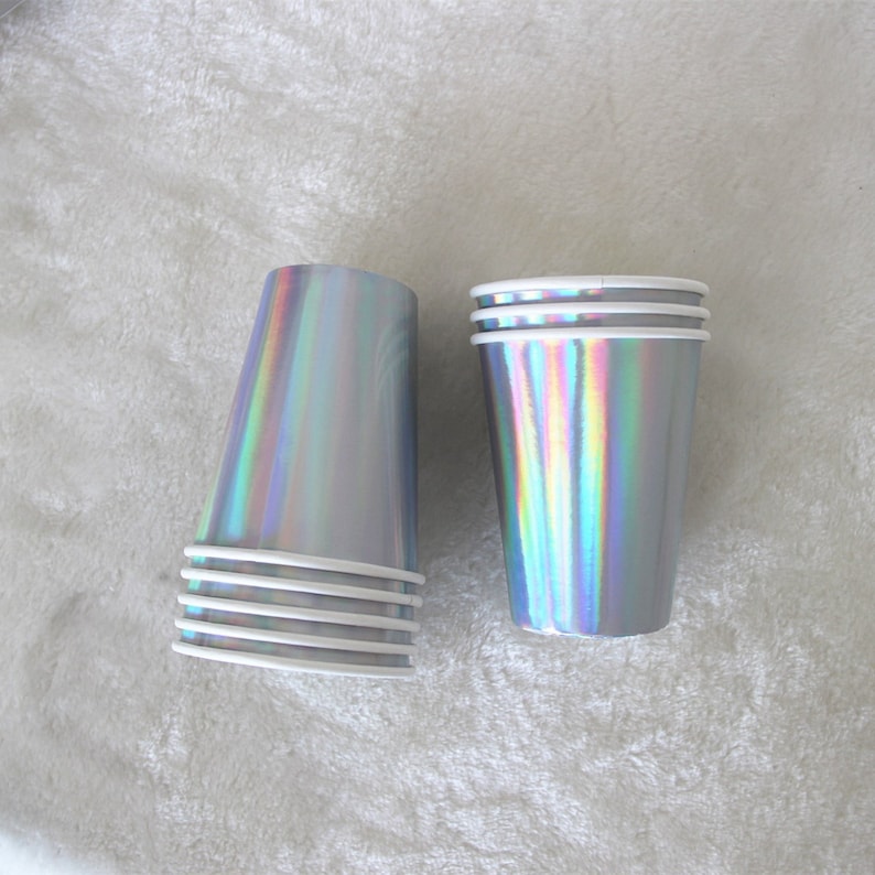 May include: A stack of iridescent silver paper cups, with one cup standing upright. The cups have a white rim and are perfect for parties or special occasions.