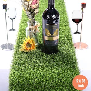 May include: A green artificial grass table runner, 12 x 36 inches, with a bottle of red wine, a wine glass, and a floral arrangement. The wine bottle has a gold label that reads "Delicious Red Wine".