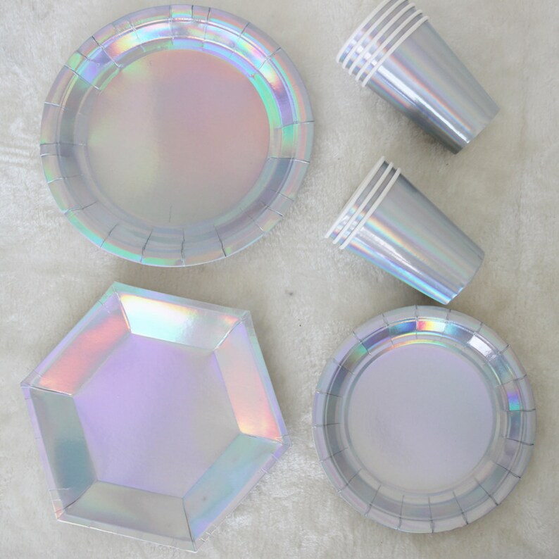 May include: A set of iridescent party supplies, including round and hexagonal paper plates and cups. The plates and cups have a holographic finish that shimmers in different colors.