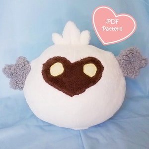 Könnte beinhalten: Ein weißes Plüschtier in Form eines Vogels mit einem braunen herzförmigen Gesicht und grauen Flügeln. Das Spielzeug sitzt auf einer hellblauen Oberfläche. Der Text '.PDF Pattern' ist in einem rosa Herz geschrieben.