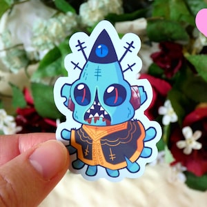May include: A blue and orange cartoon monster sticker with a single eye and stitches on its face. The monster is wearing a black and orange jacket with a cross pattern.