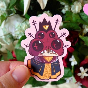 May include: A pink sticker featuring a cartoon character with four eyes, a red and black body, and a crown. The character is wearing a black and yellow robe with a cross pattern. The sticker has a pink background.