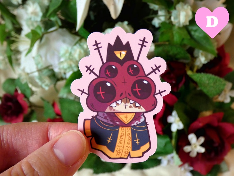 May include: A pink sticker featuring a cartoon character with four eyes, a red and black body, and a crown. The character is wearing a black and yellow robe with a cross pattern. The sticker has a pink background.