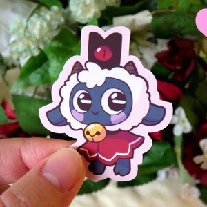May include: A sticker of a cartoon sheep wearing a red and white cape and a black crown with a red eye. The sheep has a blue face and large, white eyes.