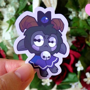 May include: A sticker featuring a cartoon black sheep with a blue crown and a purple cape. The sheep has a grumpy expression and is wearing a blue crown with a white gem.