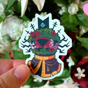 May include: A sticker featuring a cartoon creature with a green, leafy head, red flowers, and a black and orange robe. The creature has a black triangular face with a red eye and a white eye.