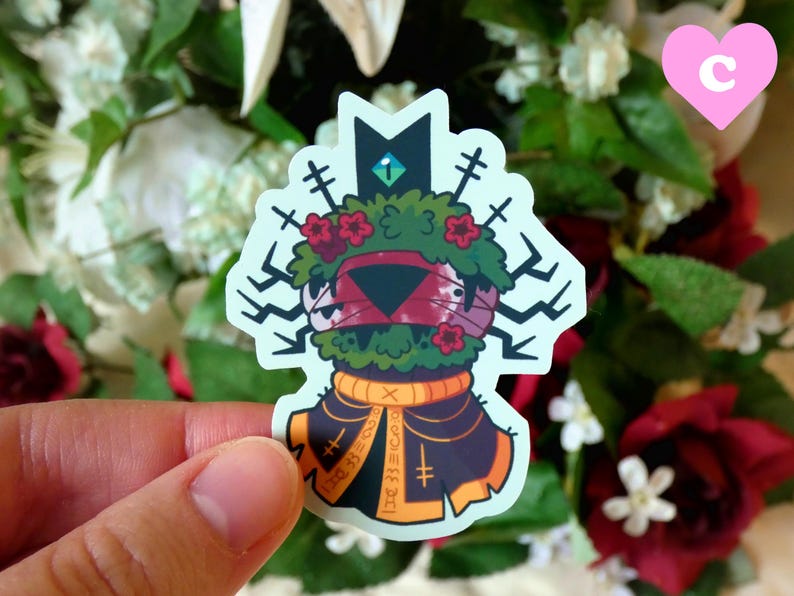 May include: A sticker featuring a cartoon creature with a green, leafy head, red flowers, and a black and orange robe. The creature has a black triangular face with a red eye and a white eye.