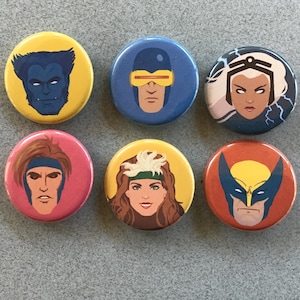 May include: A set of six circular pinback buttons featuring stylized illustrations of comic book characters. The buttons display portraits of Beast on yellow, Cyclops on blue, Storm on dark blue, Gambit on pink, Rogue on yellow, and Wolverine on orange.