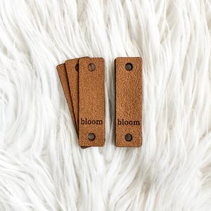 May include: Several brown leather tags with the word "bloom" engraved on them. Each tag is rectangular with rounded corners and a hole at the top and bottom. The tags are arranged on a white, fluffy surface.